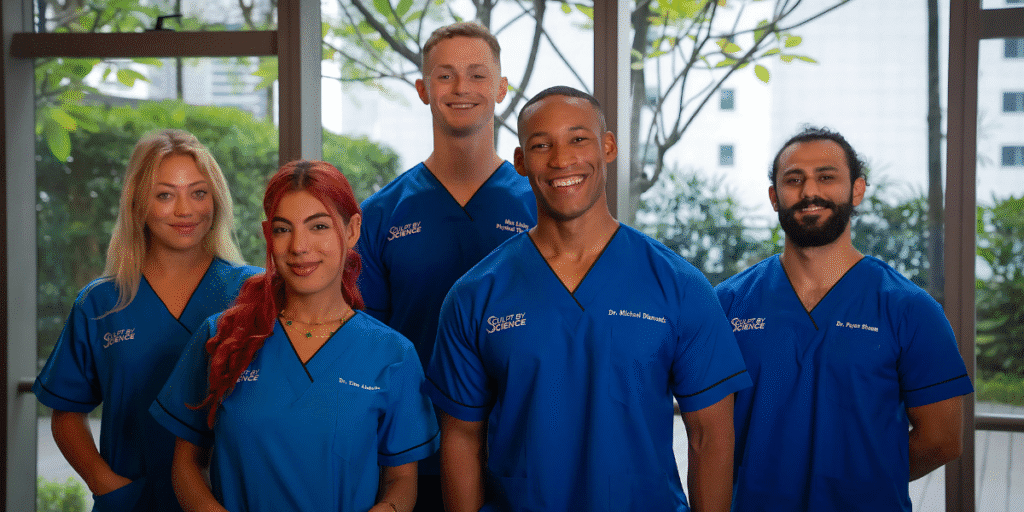 A group of five healthcare professionals wearing blue medical scrubs, standing together in a brightly lit room with plants in the background. They are smiling at the camera.