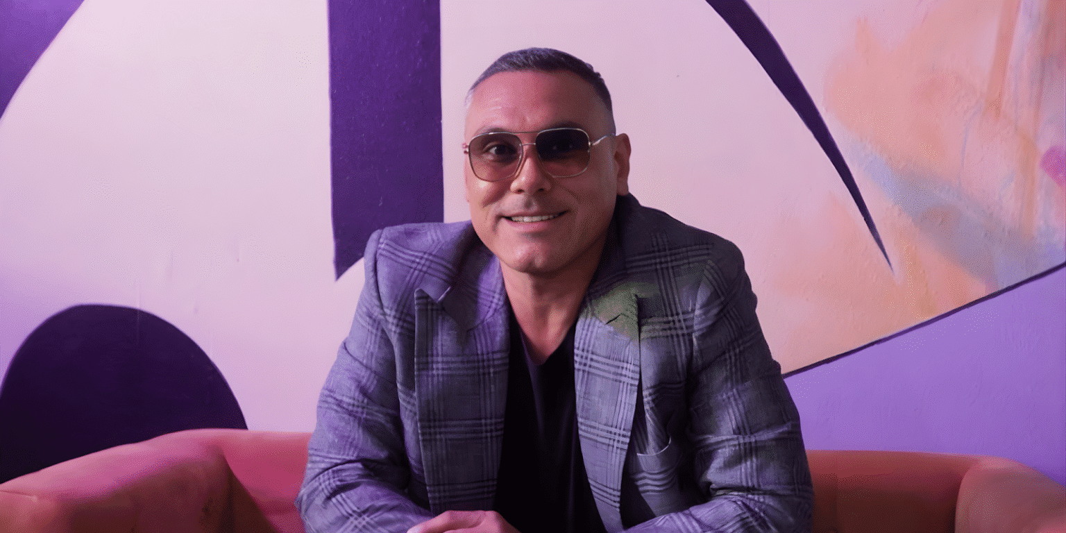 Dylan Garcia smiling in a stylish plaid jacket and sunglasses, seated in front of a colorful, artistic backdrop promoting funk music.